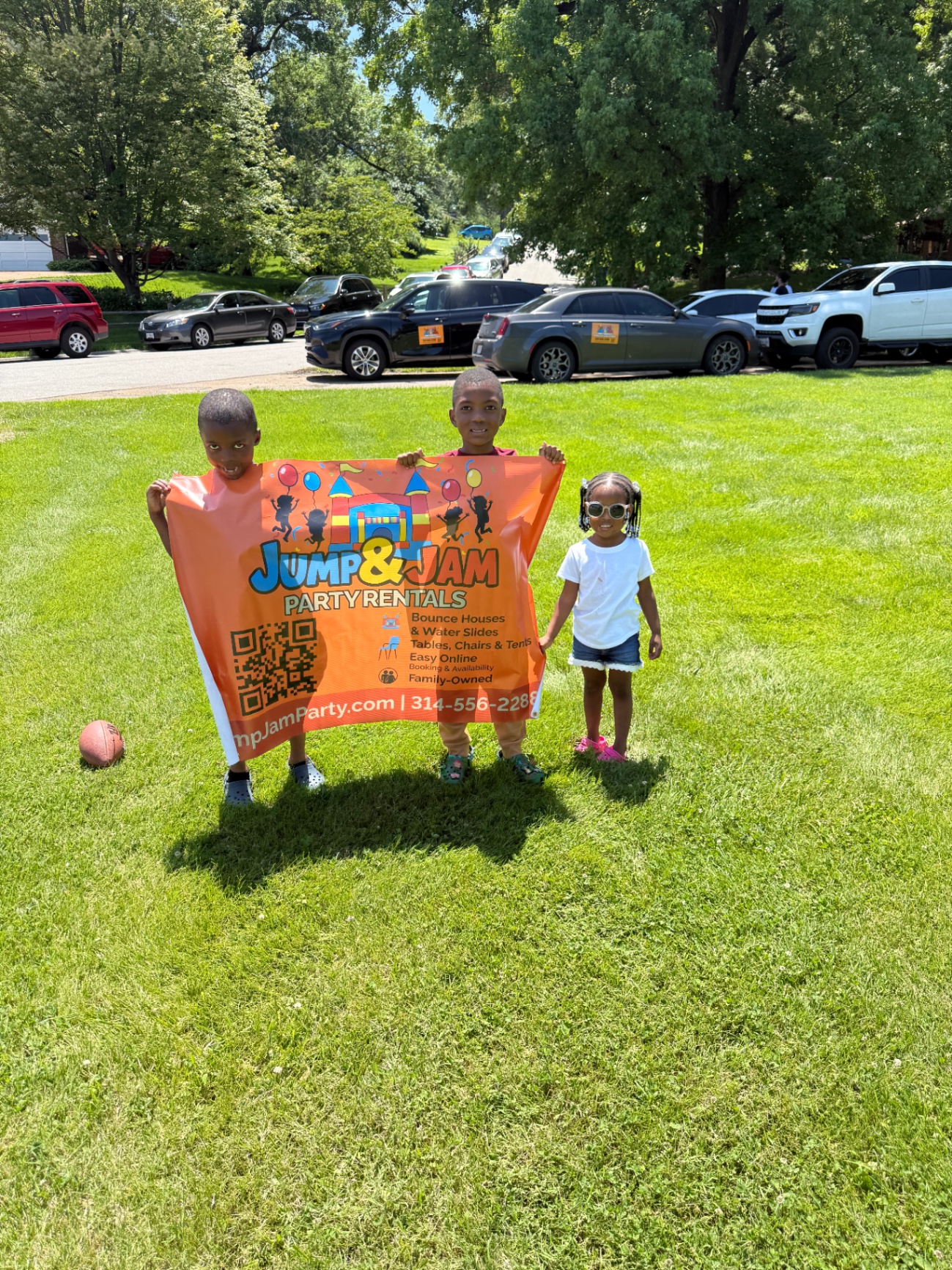 Three children holding a Jump & Jam Party Rentals banner on the grass at a community event